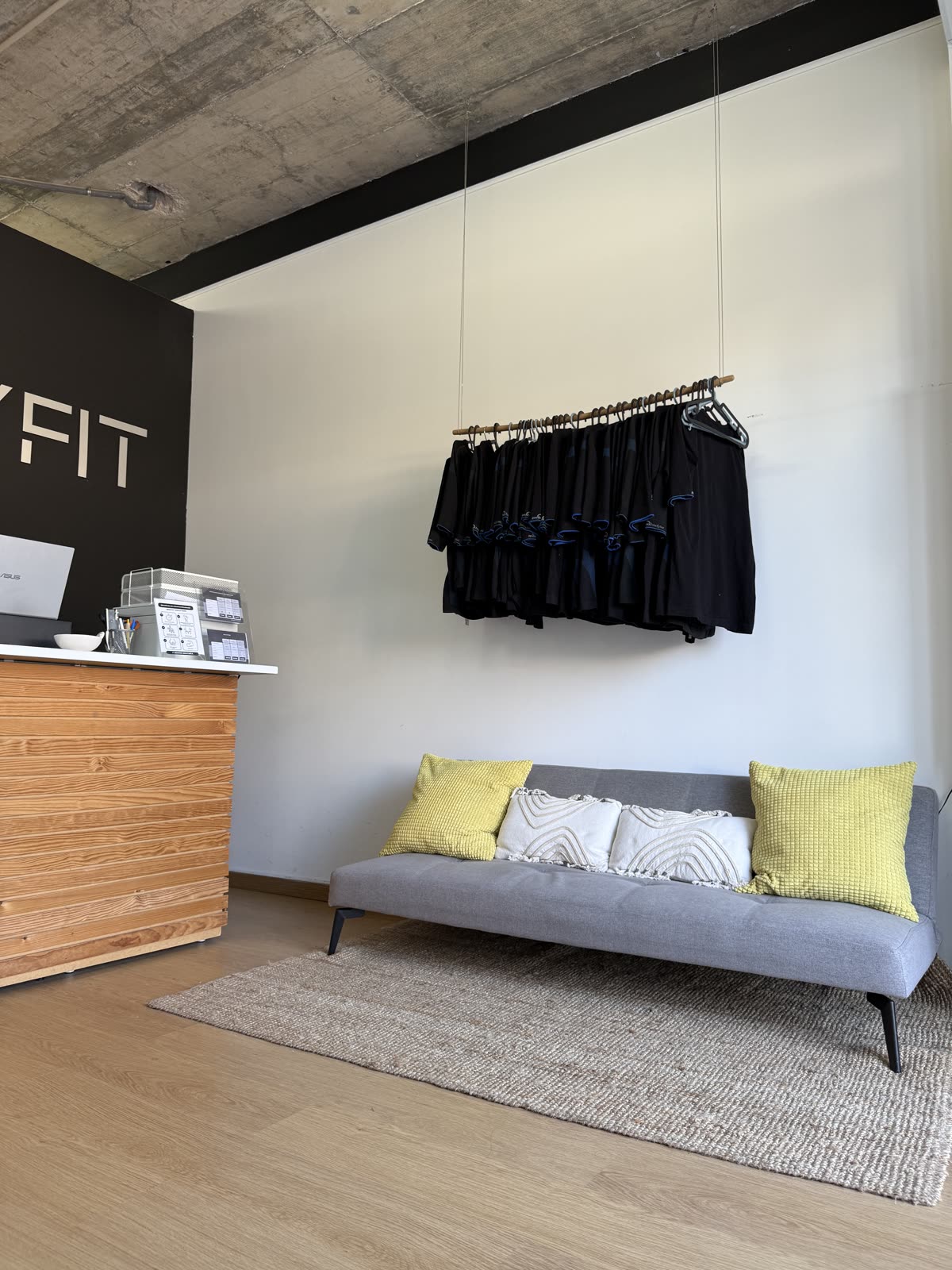 Zona de receção e lounge do BODYFIT com sofá cinzento, almofadas amarelas e roupa da marca.